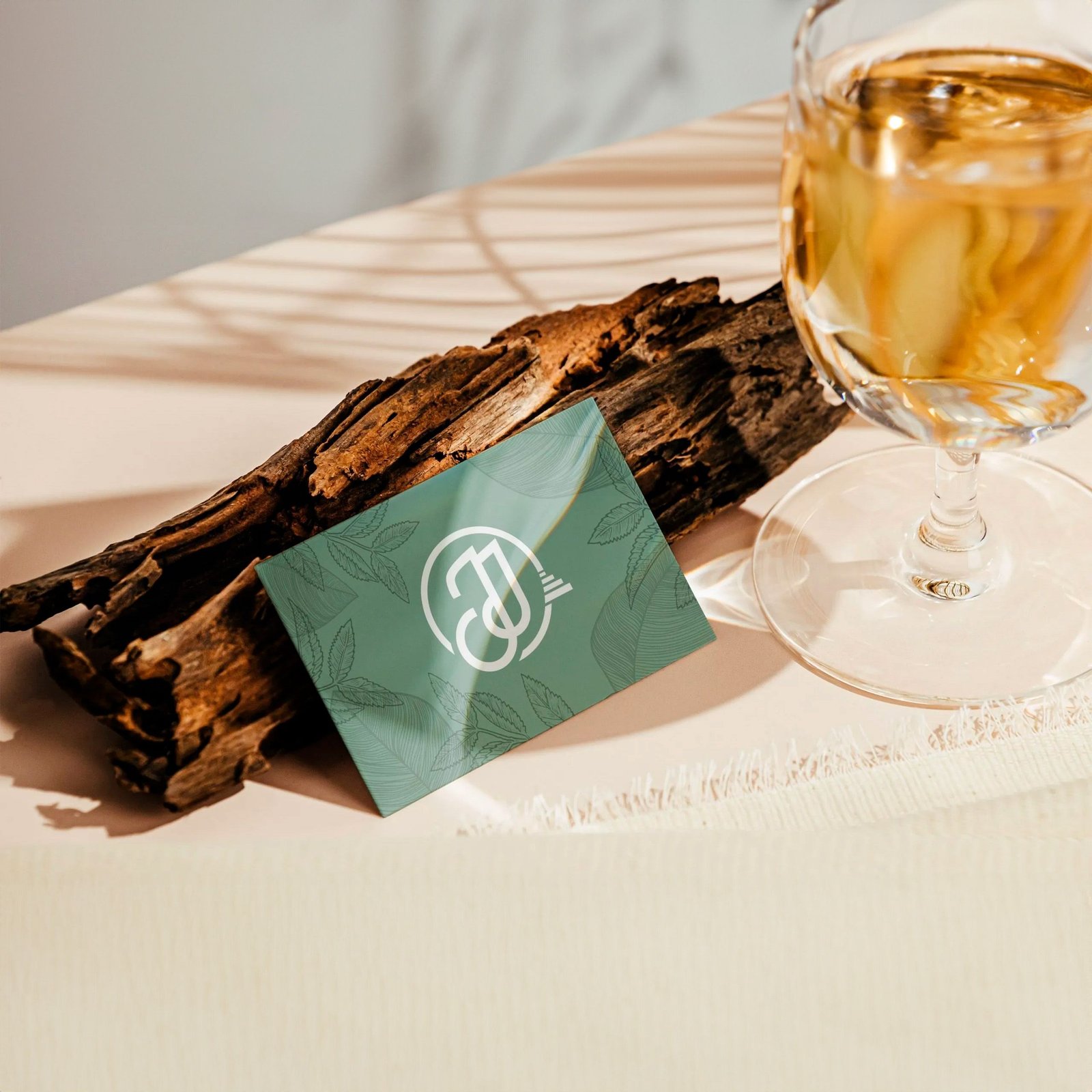 Una copa de vino blanco reposa sobre una mesa junto a un trozo de madera flotante y una tarjeta verde con un logotipo. El fondo está tenuemente iluminado y proyecta sombras sutiles.