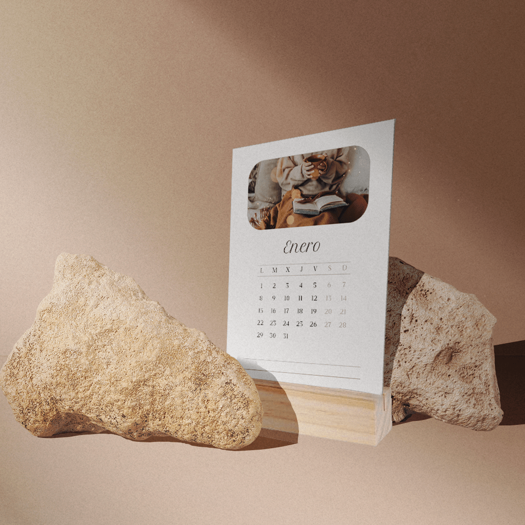 El "Calendario personalizable" de enero, con la etiqueta "Enero", se muestra entre dos rocas texturizadas sobre una base de madera. Una pequeña foto aparece sobre el nombre del mes. Una suave luz natural ilumina la escena, creando sombras sutiles.