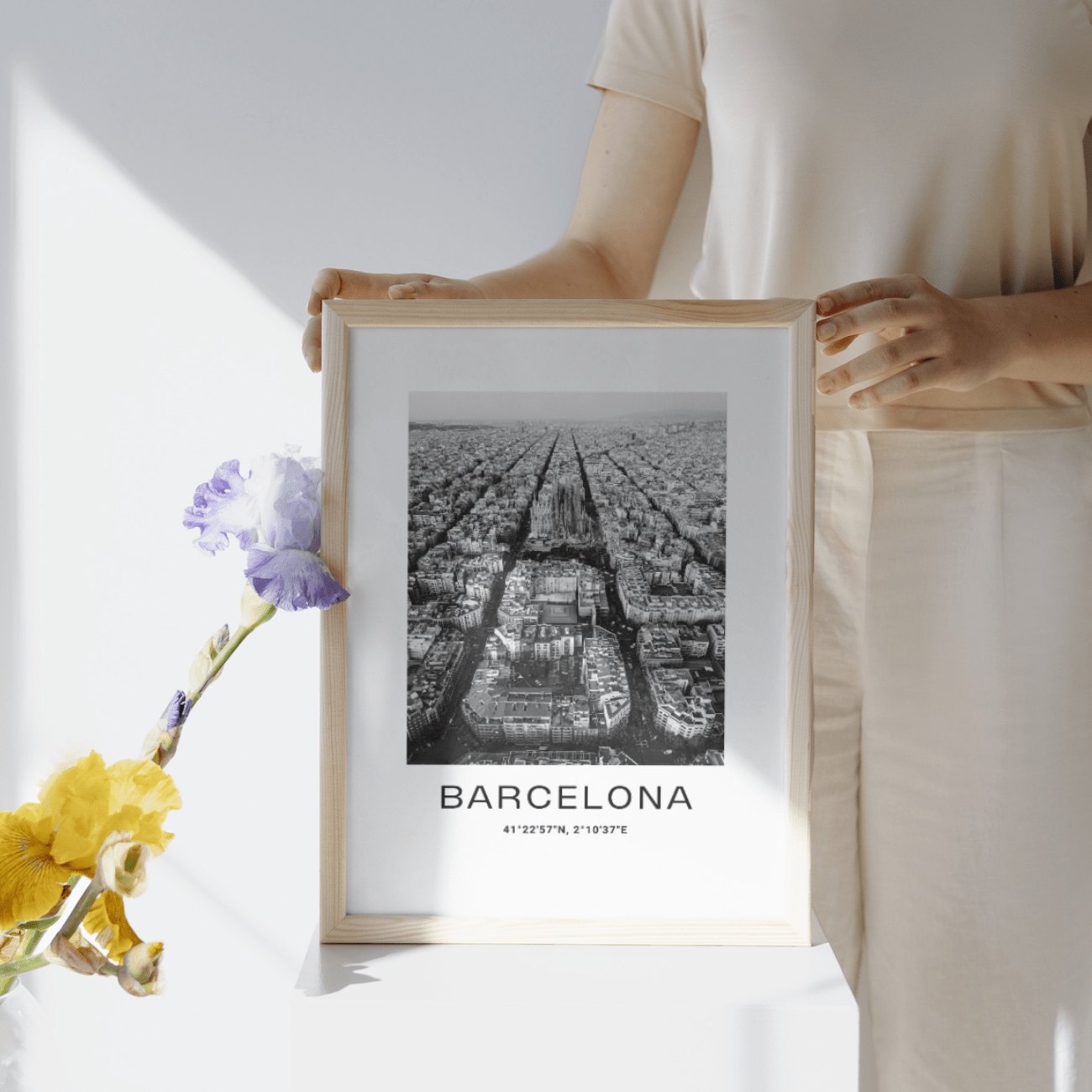 Alguien sostiene el "Póster Barcelona A3", una lámina en blanco y negro de un paisaje urbano con coordenadas. Está expuesta en una habitación soleada junto a un jarrón con flores de colores.