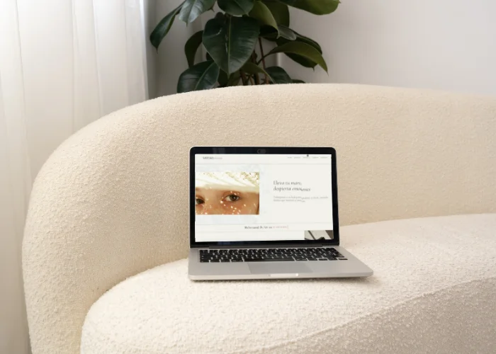 Una laptop con una página web y la imagen de un ojo se encuentra sobre un sofá color crema. Al fondo, cerca de una cortina, se ve una planta verde. El ambiente se ve tranquilo y minimalista.